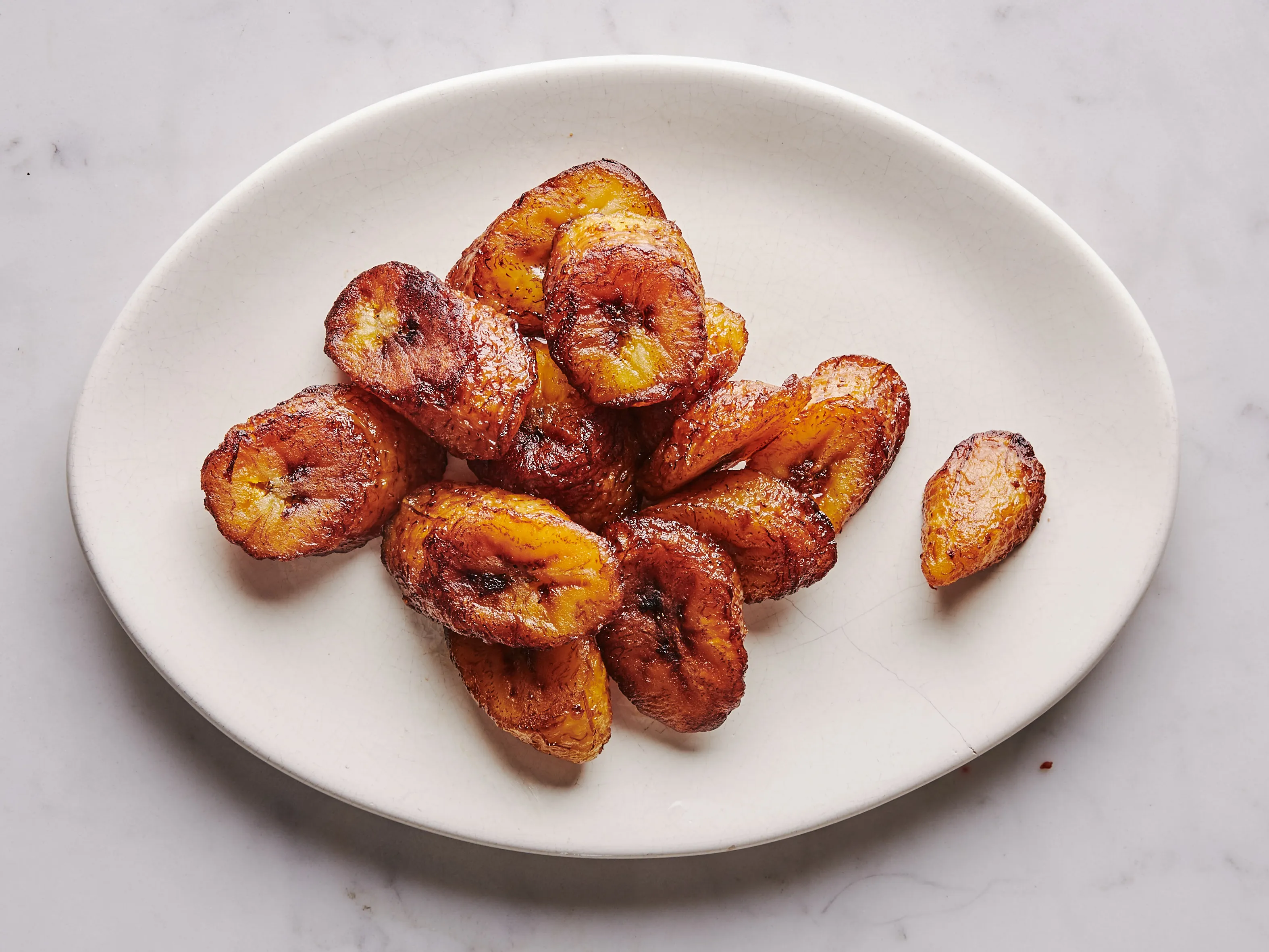 A white plate of golden fried sweet plantains (maduros) with caramelized edges on a wooden table setting, warm natural kitchen light