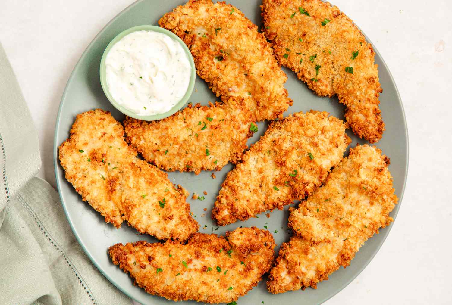 A white plate piled with crispy golden air fryer chicken tenders with a small bowl of honey mustard on a wooden table in a bright home kitchen
