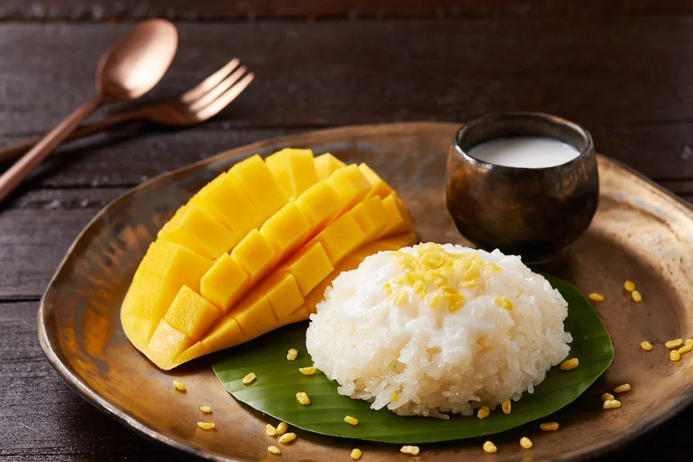 A white plate with a neat mound of coconut sticky rice beside sliced ripe mango, drizzled with coconut sauce and sprinkled with toasted sesame seeds, set on a warm wooden table in soft natural light