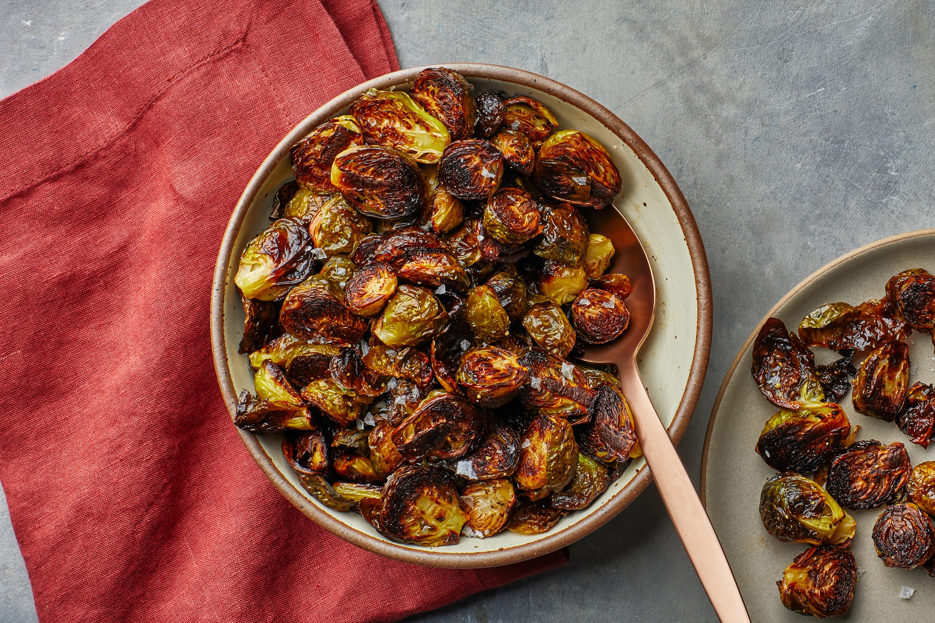 A white serving bowl filled with deeply golden roasted Brussels sprouts with crisp edges, lightly glossy from lemon and butter, on a warm kitchen table with a neutral linen napkin