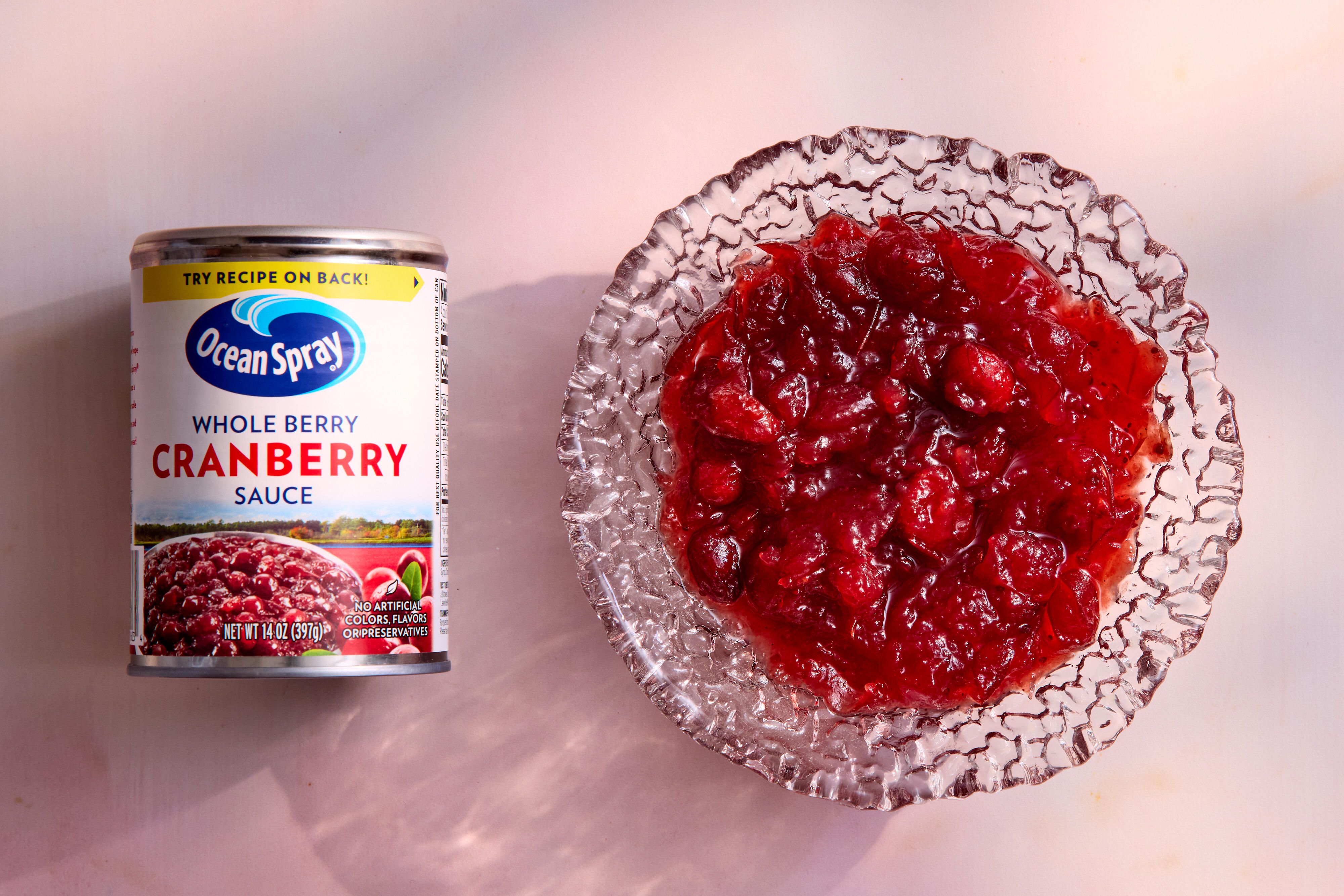 A white serving bowl filled with glossy deep-red homemade cranberry sauce with visible whole cranberries on a wooden table, warm kitchen lighting