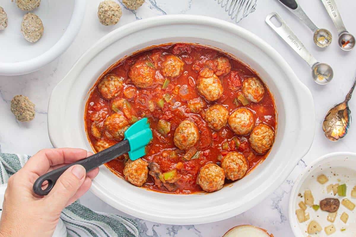 A white serving bowl filled with saucy Crock-Pot meatballs in a rich tomato marinara, topped with chopped parsley, on a warm wood table with a serving spoon