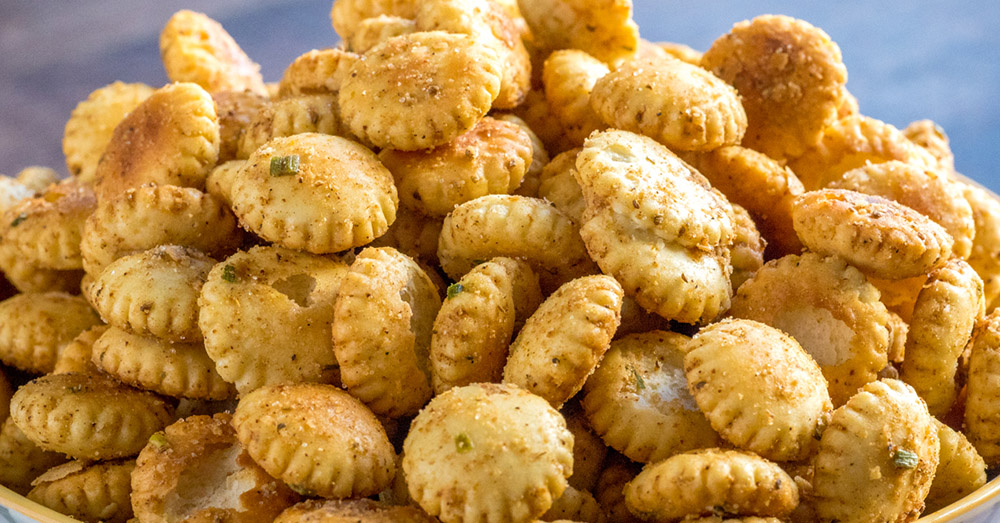 A white serving bowl piled high with spicy ranch fire crackers made from oyster crackers, lightly glossy with seasoning, on a warm kitchen table with a small linen napkin