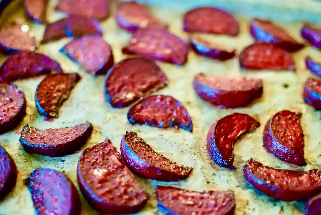 A white serving platter piled with glossy roasted beetroot wedges, sprinkled with flaky salt and fresh herbs on a warm-toned kitchen table