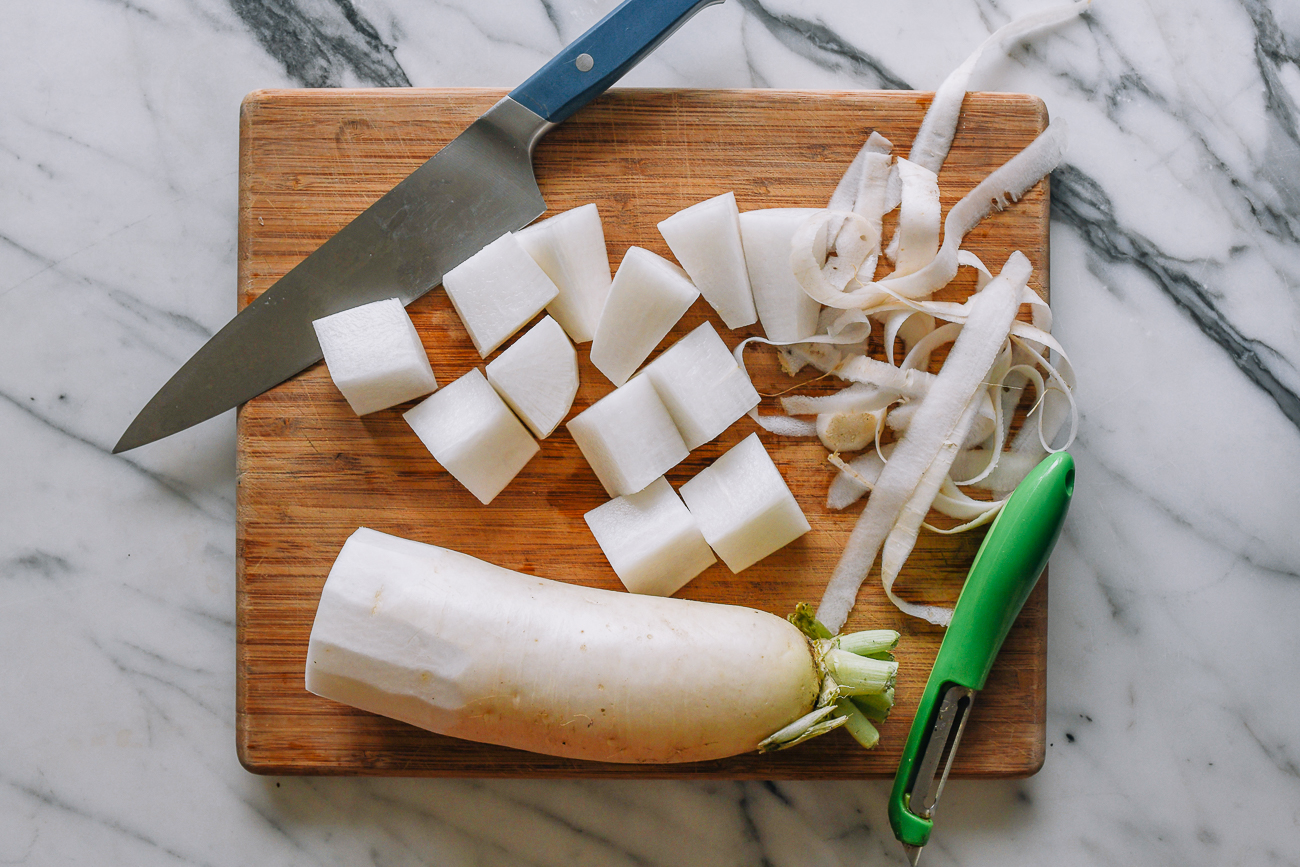 A whole daikon radish and a peeled section on a wooden cutting board with a chef's knife in a home kitchen prep setup