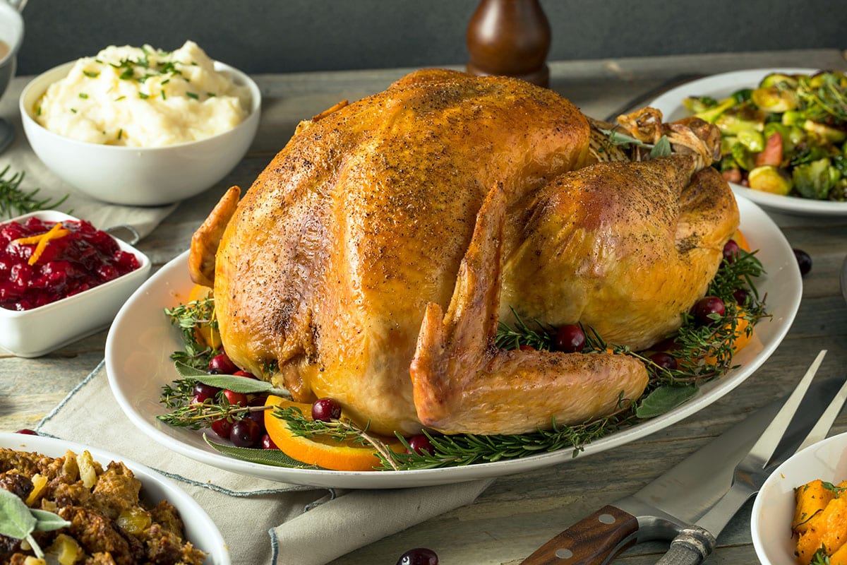 A whole golden-brown roast Thanksgiving turkey resting on a white platter with fresh herbs and roasted citrus on a wooden dining table in warm natural kitchen lighting