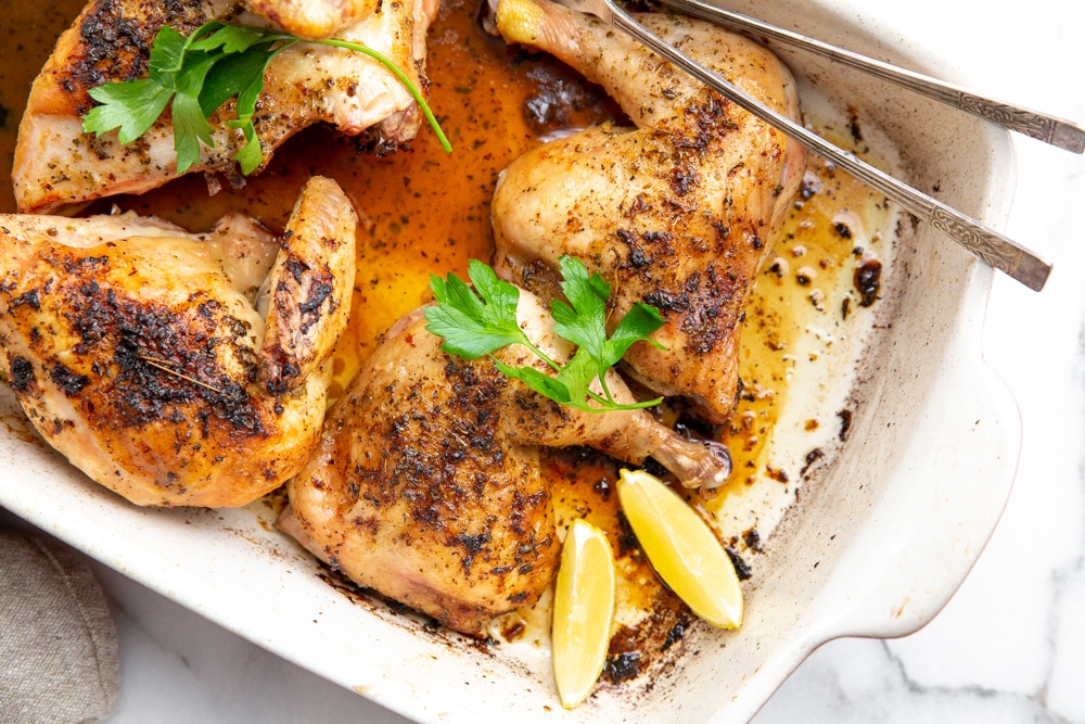 A whole lemon-garlic roast chicken in a skillet with golden crispy skin, surrounded by roasted shallots and lemon halves on a warm kitchen counter