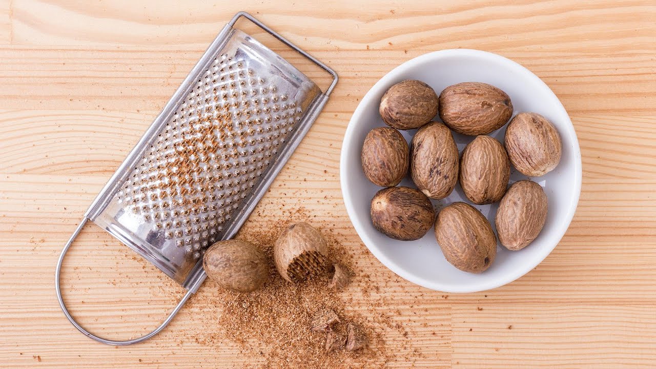 A whole nutmeg and a microplane grater resting on a cutting board on a home kitchen counter with soft warm lighting