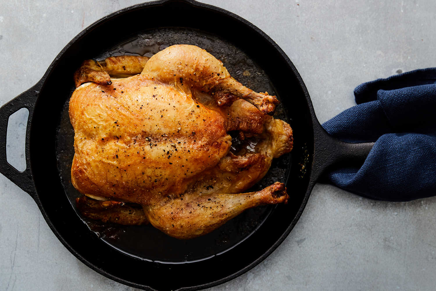 A whole oven roasted chicken with deeply golden, crisp skin resting in a cast iron skillet with lemon halves and herbs on a warm kitchen counter