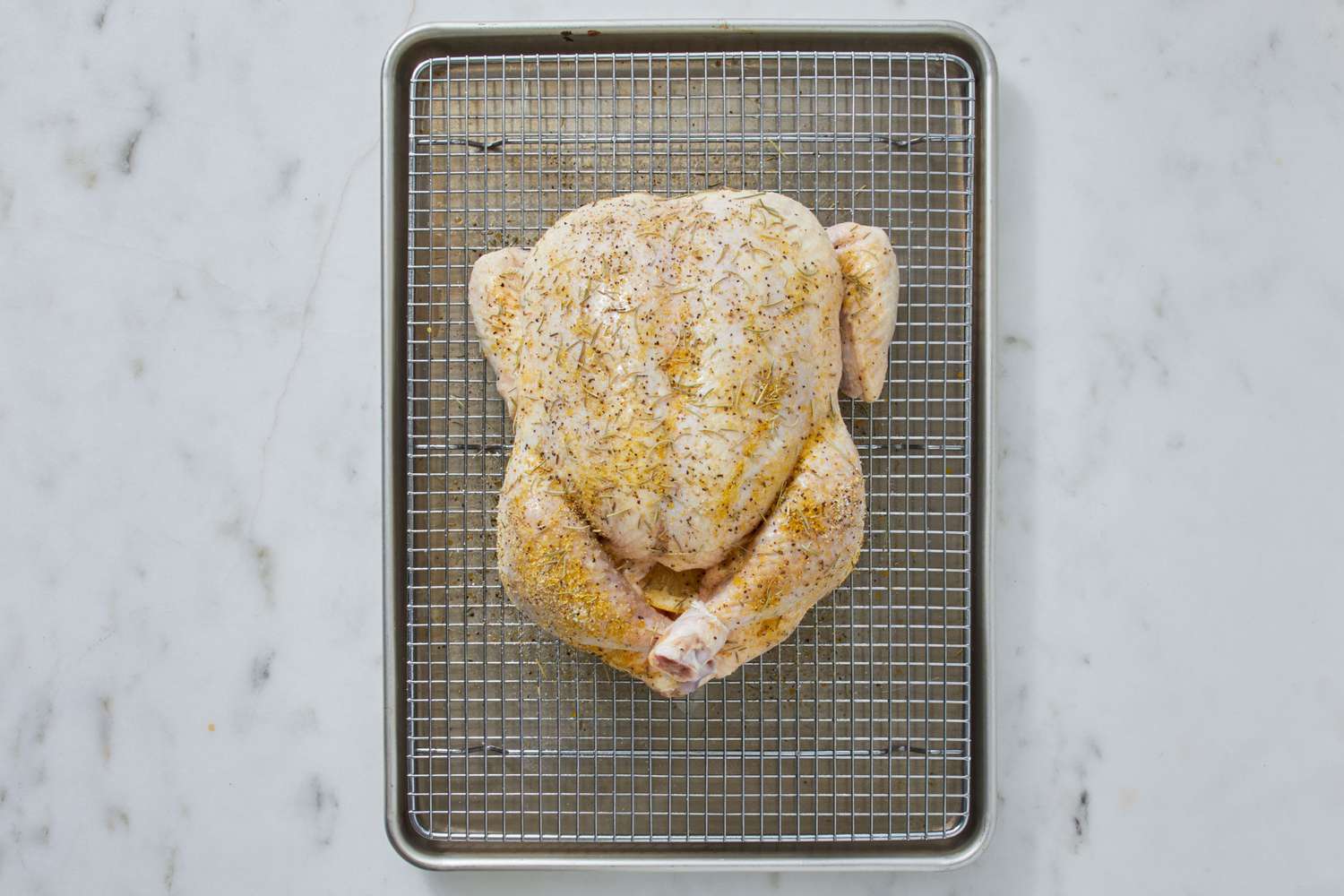 A whole raw chicken seasoned and sitting on a wire rack over a sheet pan, ready to roast