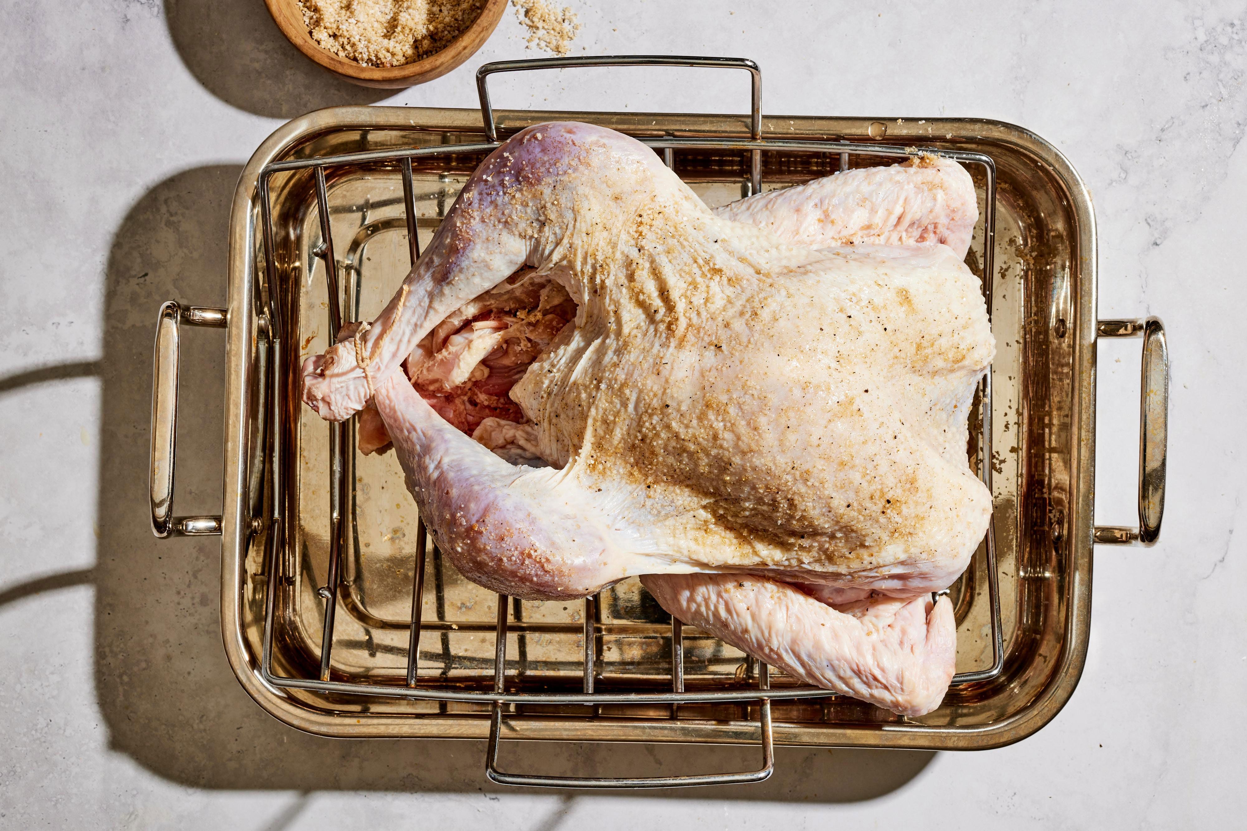 A whole raw turkey on a wire rack over a sheet pan, lightly seasoned with salt and pepper, sitting uncovered in a refrigerator