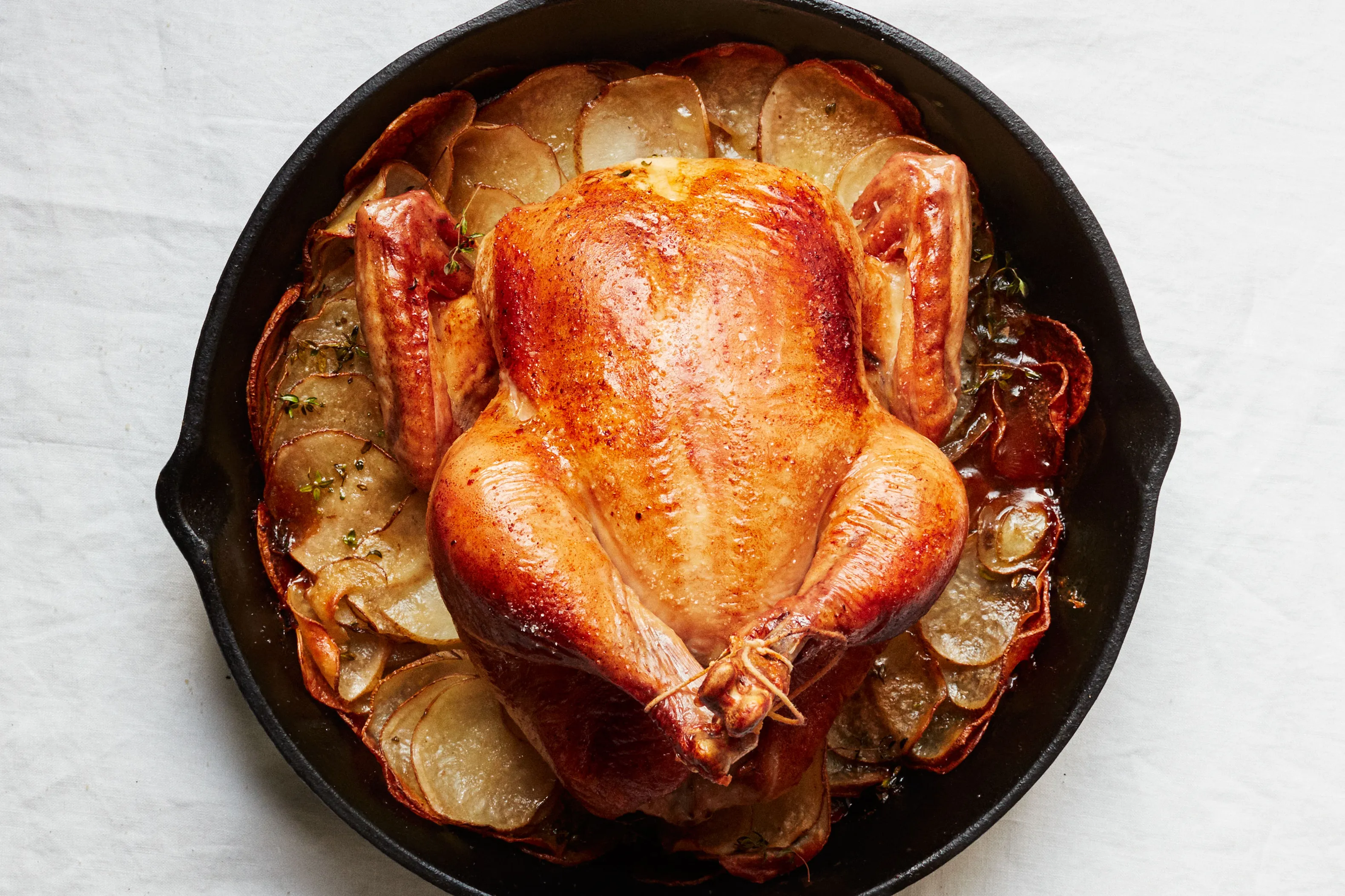 A whole roasted chicken with deeply golden, crisp skin resting in a cast iron skillet with roasted lemon halves and thyme sprigs on a warm kitchen counter