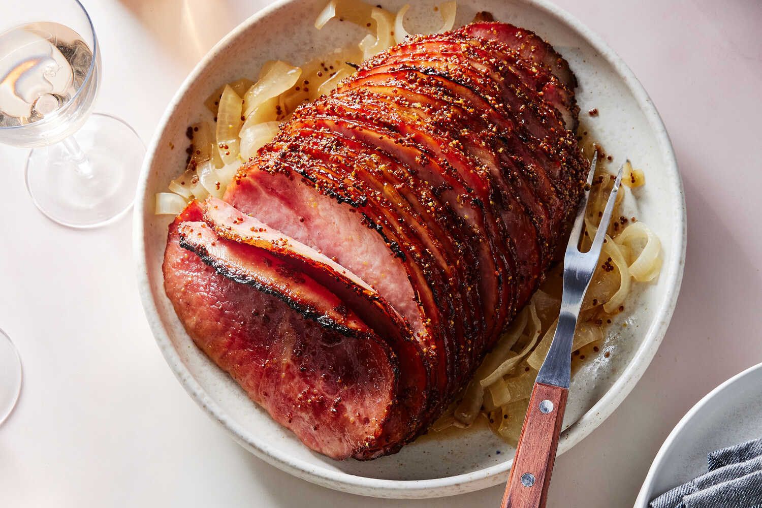 A whole spiral-sliced ham on a white platter with a glossy honey glaze and caramelized edges, surrounded by rosemary and orange slices on a warm wooden table