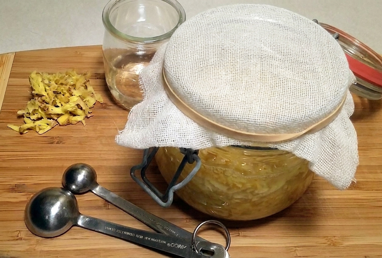 A wide-mouth glass jar covered with a cloth and rubber band on a kitchen counter, with grated ginger and a spoon nearby