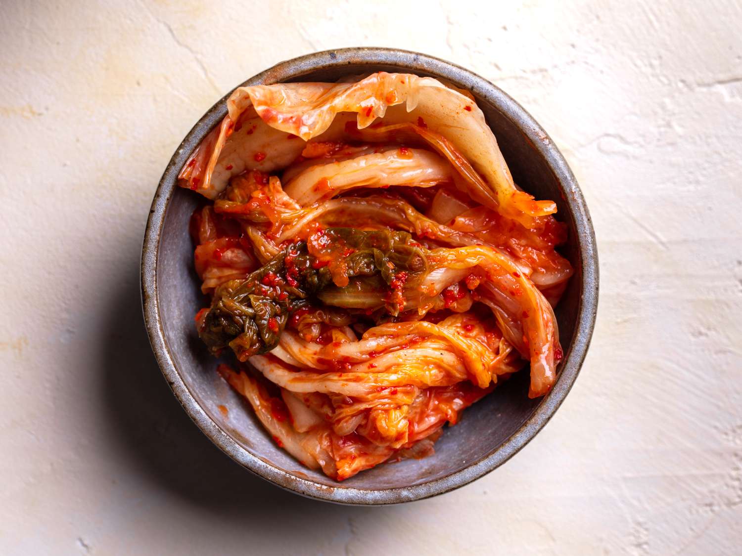 A wide-mouth glass jar packed with bright red napa cabbage kimchi on a kitchen counter, with a small bowl of gochugaru and garlic nearby
