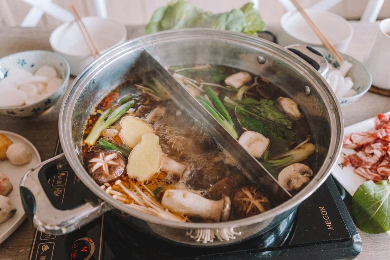 A wide pot of hot pot broth simmering with ginger slices, scallions, and mushrooms on a stovetop