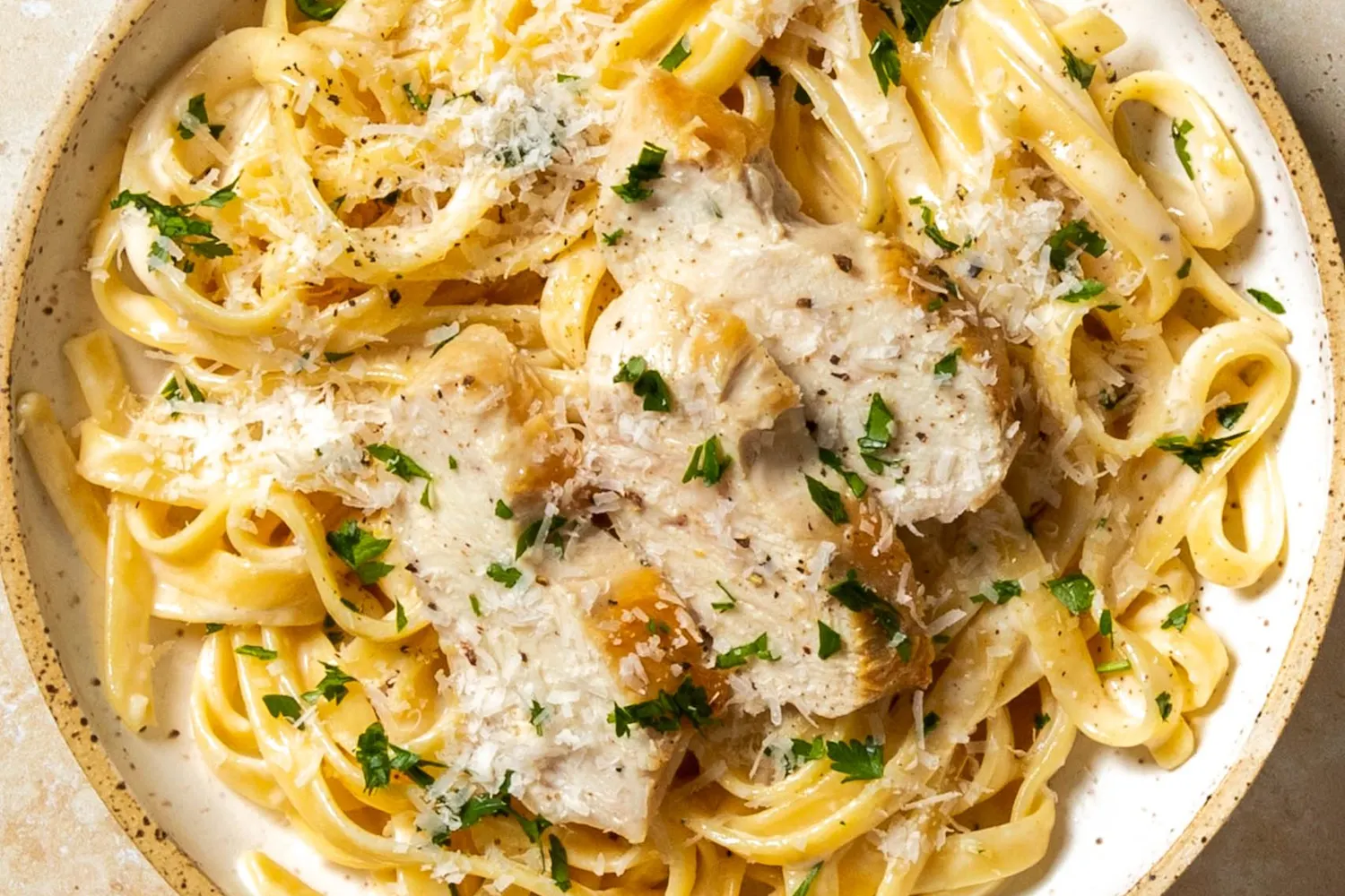 A wide shallow bowl of chicken Alfredo pasta with fettuccine coated in glossy Parmesan cream sauce, sliced seared chicken on top, and a sprinkle of black pepper on a simple dinner table
