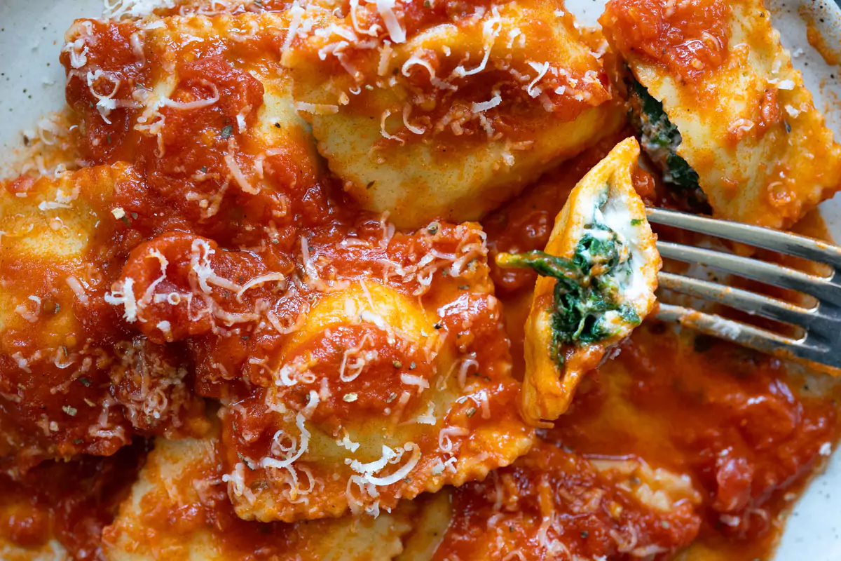 A wooden board of freshly filled ravioli with a piping bag nearby and a bowl of ricotta spinach filling on a bright kitchen counter