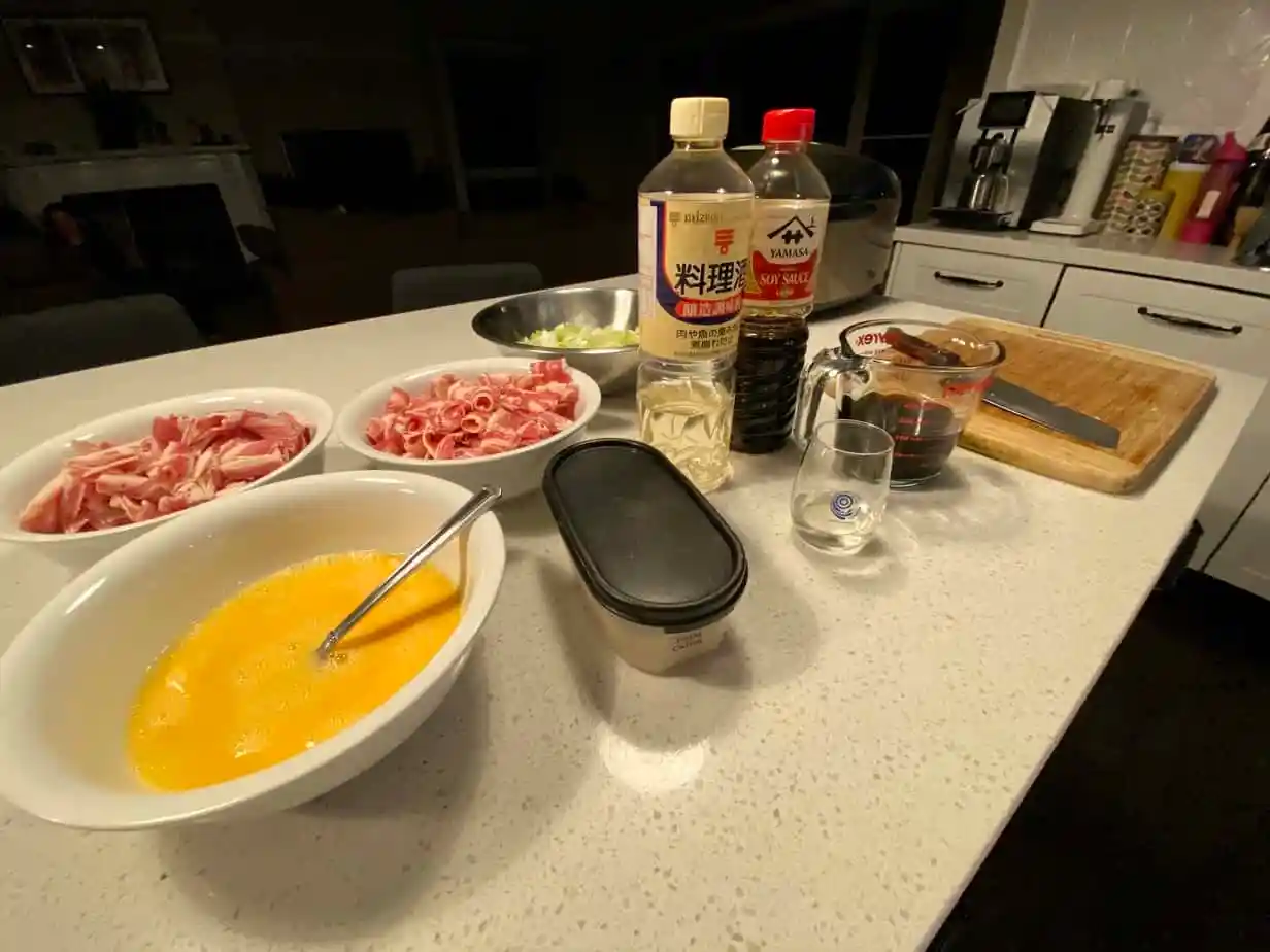 A wooden cutting board and bowls holding thin sliced beef, napa cabbage, shiitake mushrooms, tofu, scallions, and udon noodles arranged on a warm kitchen counter