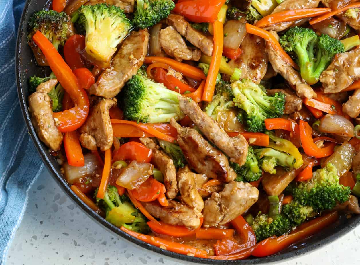 A wooden cutting board on a kitchen counter with sliced pork, soy sauce, cornstarch, garlic, ginger, and bowls of prepped vegetables ready for stir fry
