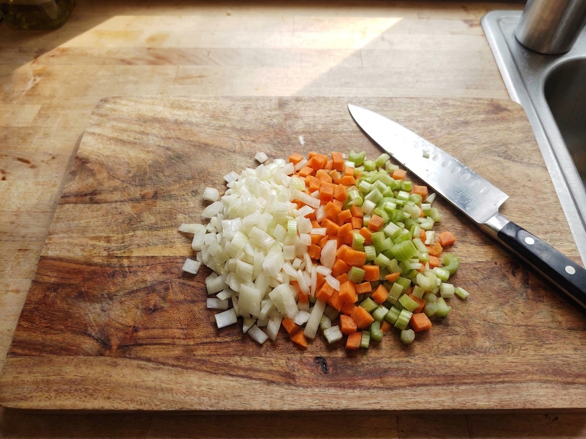 A wooden cutting board on a warm-lit kitchen counter with neatly diced onions, carrots, and celery arranged in small piles next to a chef’s knife