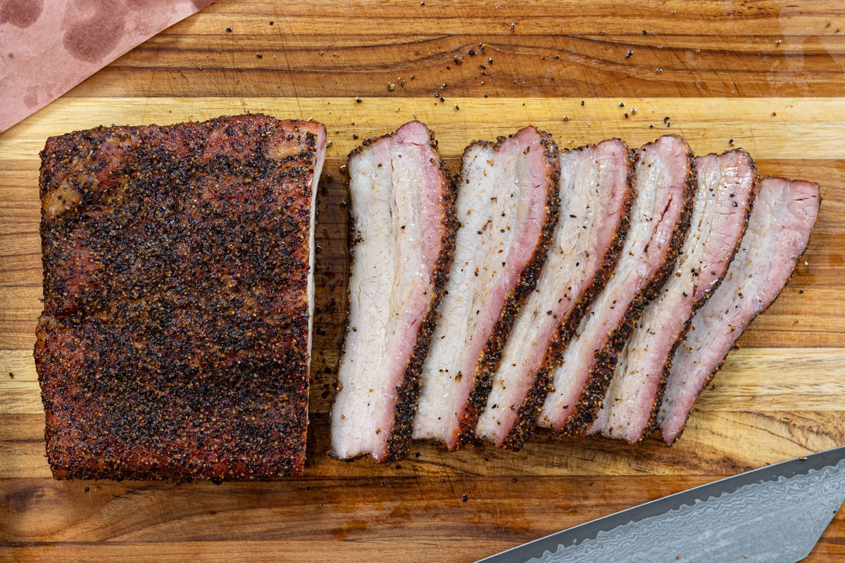 A wooden cutting board with thick slices of smoked pork belly showing a dark bark, pink smoke ring, and glossy rendered fat, set on a warm kitchen counter