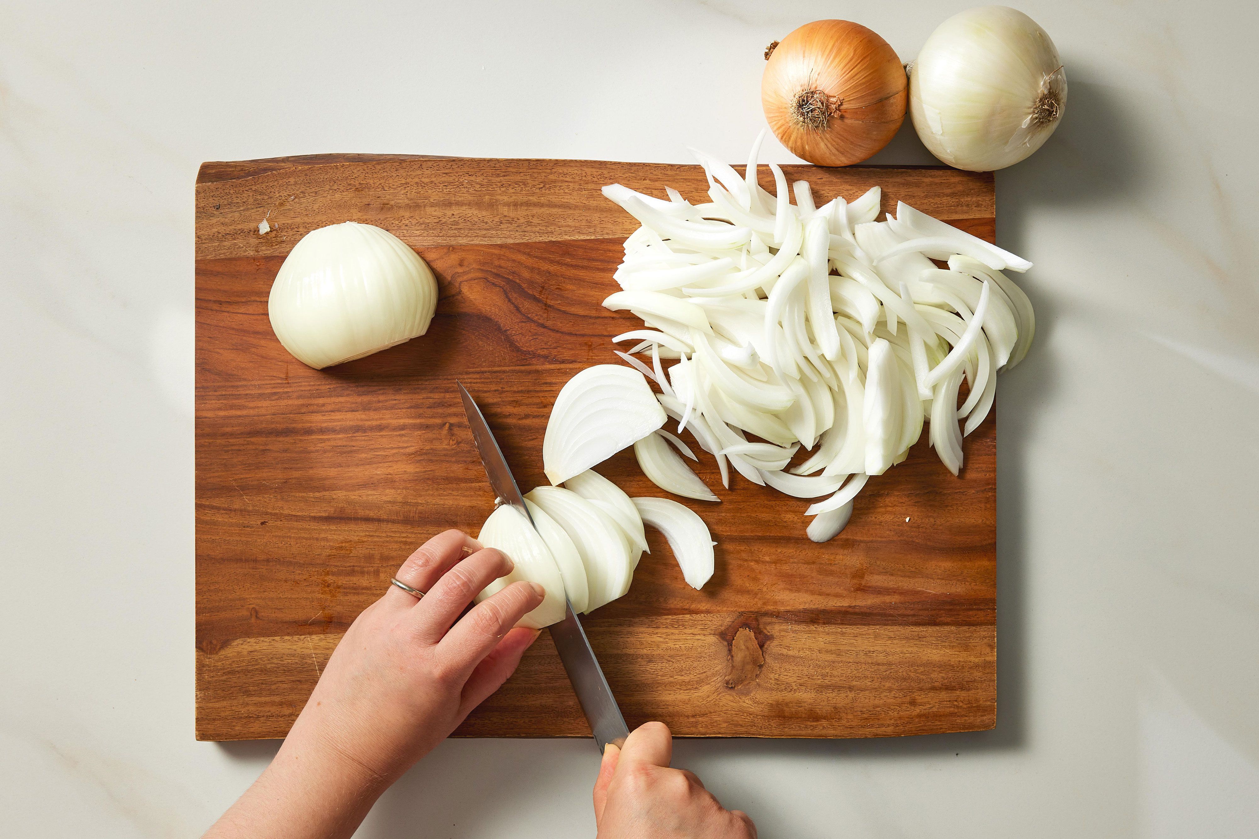 A wooden cutting board with thinly sliced yellow onions beside a small bowl of flour and spices on a bright kitchen counter