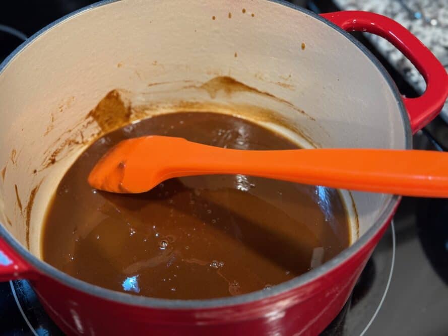 A wooden spoon stirring a dark roux in a heavy Dutch oven on a stovetop, showing glossy brown color