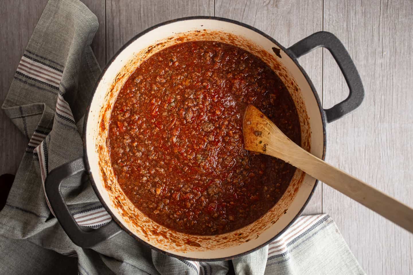 A wooden spoon stirring a simmering tomato meat sauce in a large skillet on a stovetop