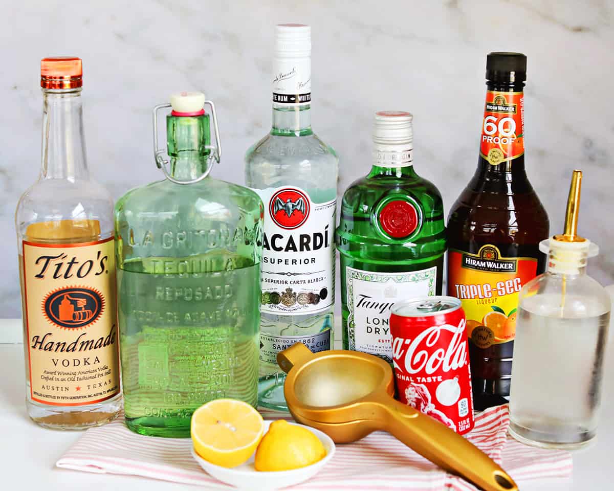 All Long Island Iced Tea ingredients arranged on a kitchen counter: vodka, gin, white rum, tequila, triple sec, fresh lemons, simple syrup, cola, and a tall glass of ice