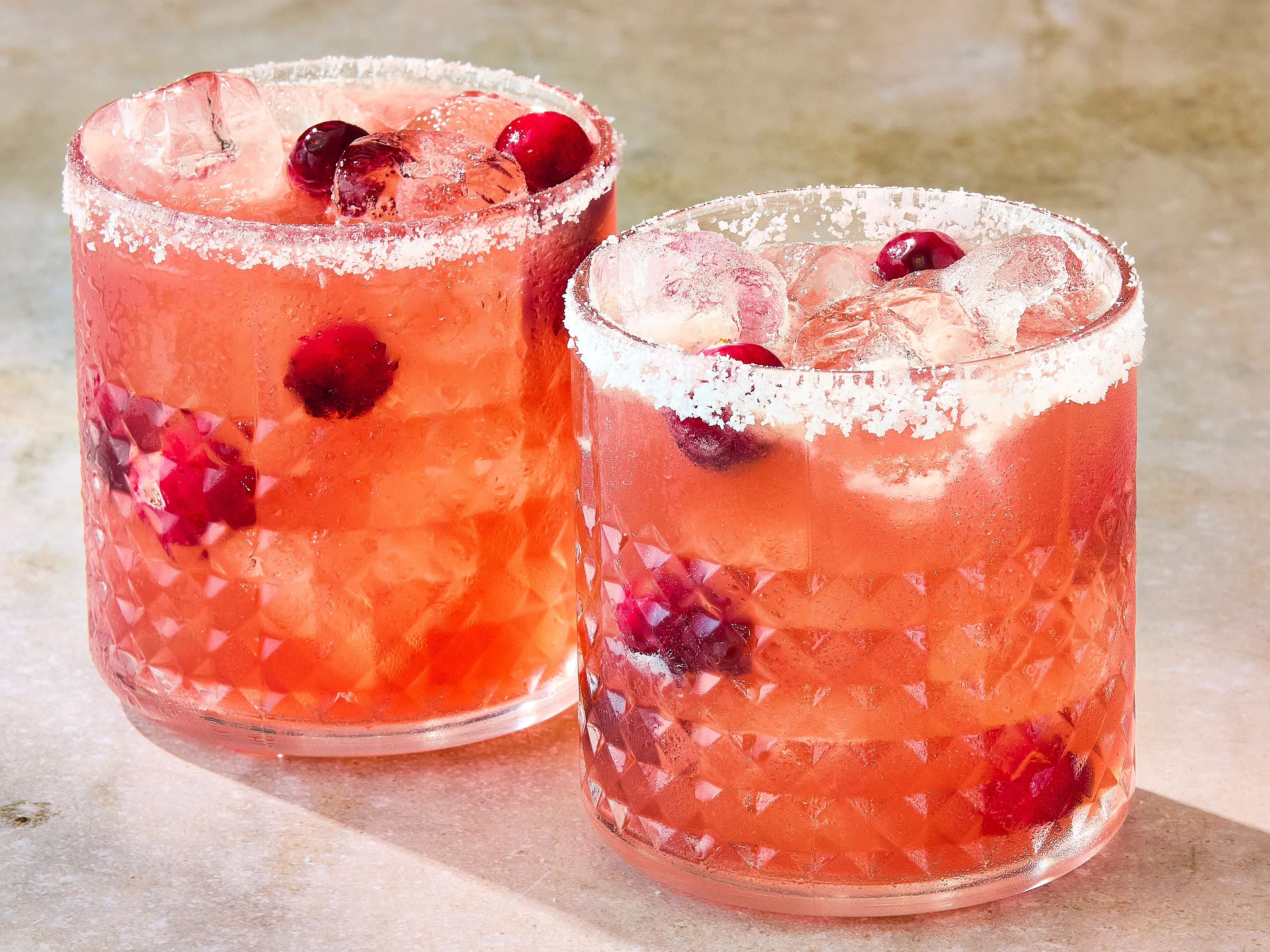 All cranberry margarita ingredients arranged on a kitchen counter, including tequila, orange liqueur, unsweetened cranberry juice, fresh limes, ice, salt, sugar, and fresh cranberries