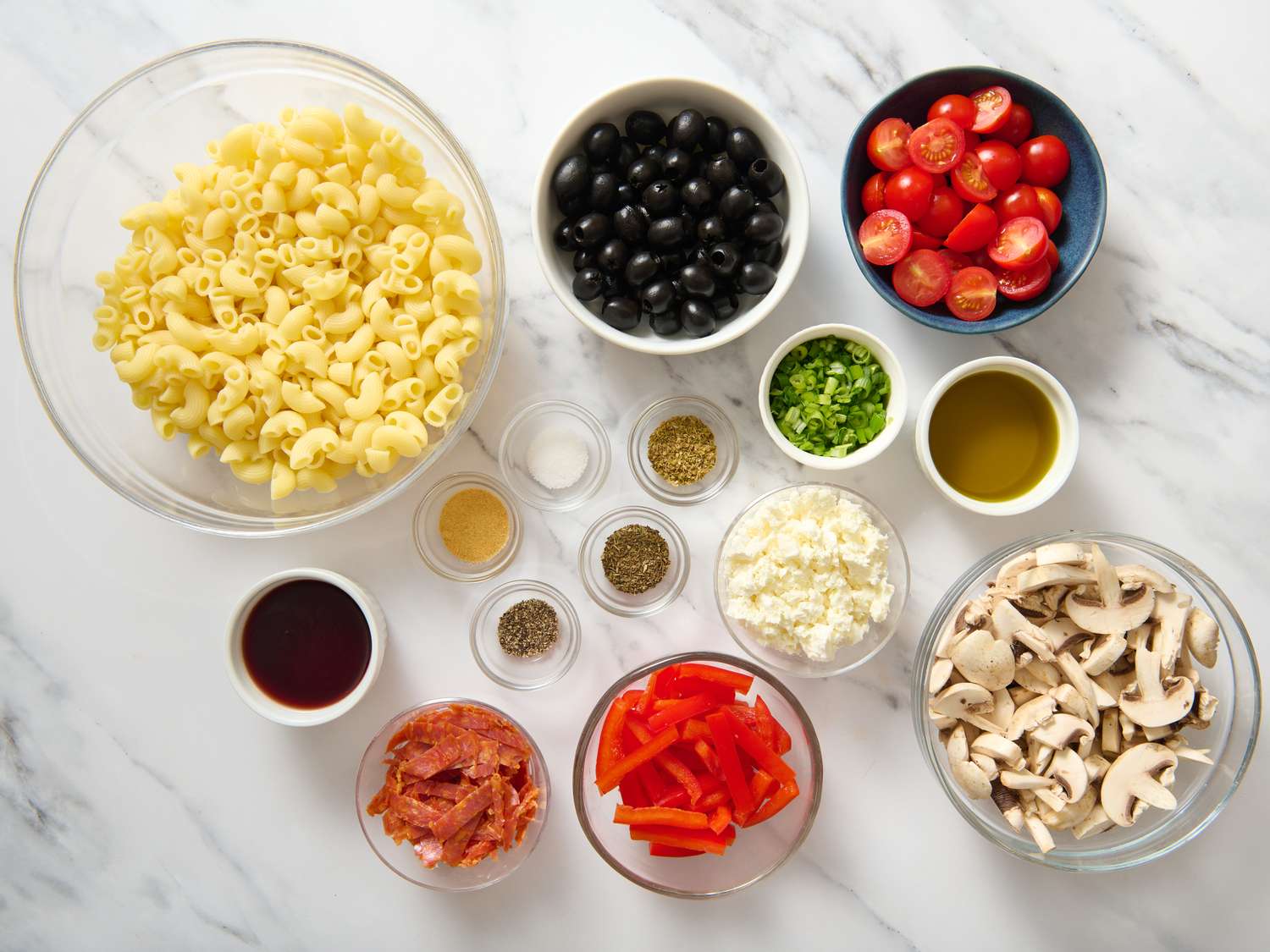 All ingredients for Greek pasta salad arranged on a light kitchen counter including rotini pasta, cucumbers, cherry tomatoes, red onion, bell pepper, feta, kalamata olives, lemon, olive oil, and dried oregano
