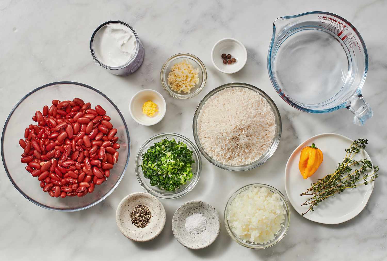All ingredients for Jamaican rice and peas arranged on a kitchen counter including long grain rice, canned kidney beans, coconut milk, scallions, thyme, garlic, onion, and Scotch bonnet pepper