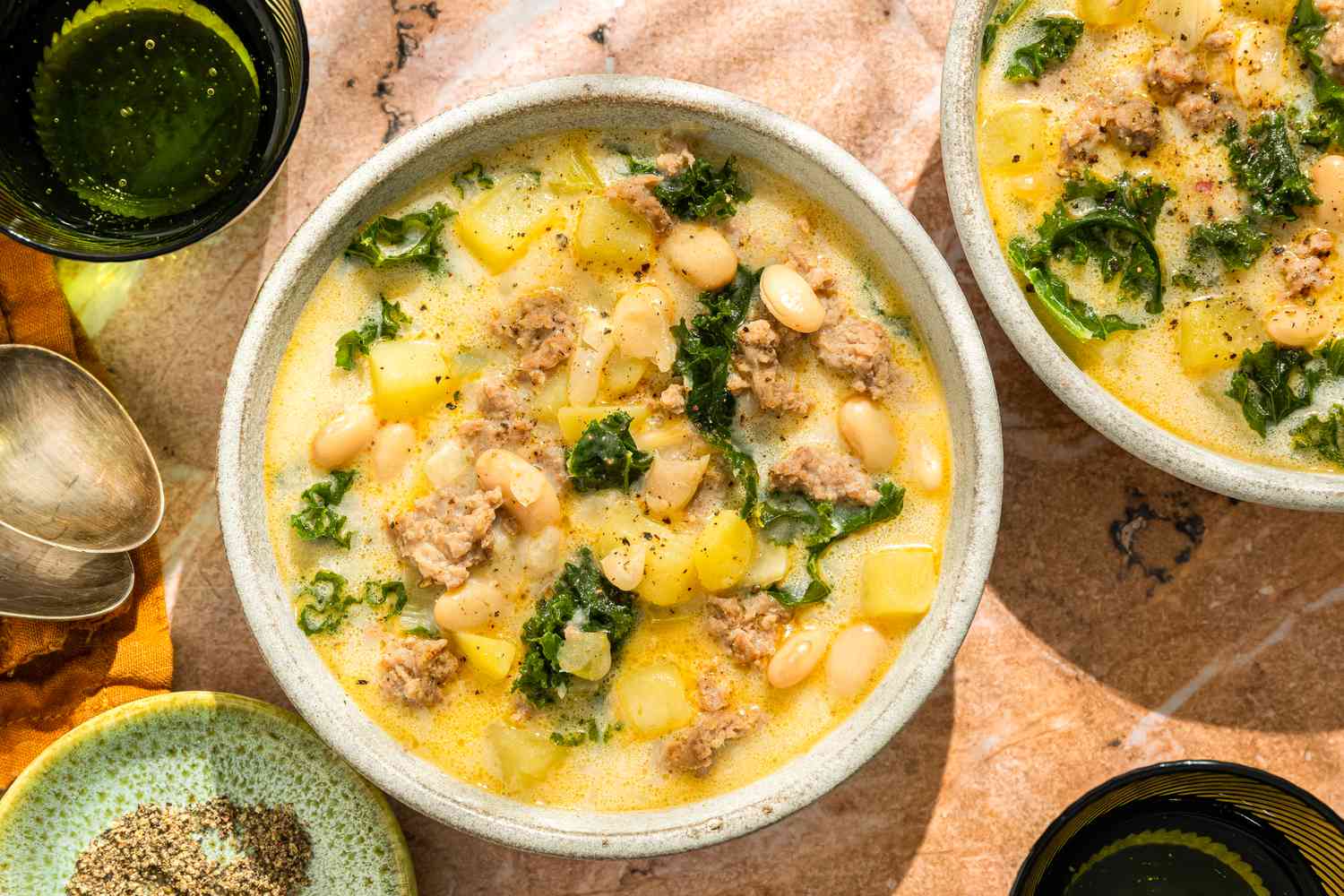 All ingredients for Zuppa Toscana soup arranged on a clean kitchen counter, including Italian sausage, potatoes, kale, onion, garlic, chicken broth, cream, and bacon
