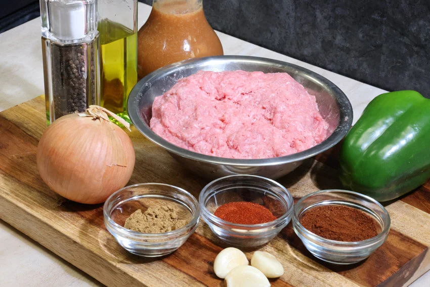 All ingredients for beanless chili arranged on a light kitchen counter including ground beef, onions, peppers, spices, tomato paste, crushed tomatoes, and broth