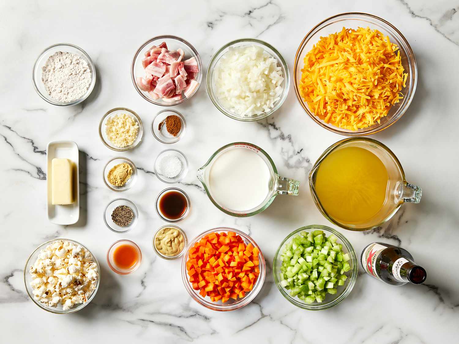 All ingredients for beer cheese soup arranged on a tidy kitchen counter, including cheddar, beer, broth, onions, carrots, celery, bacon, flour, and seasonings