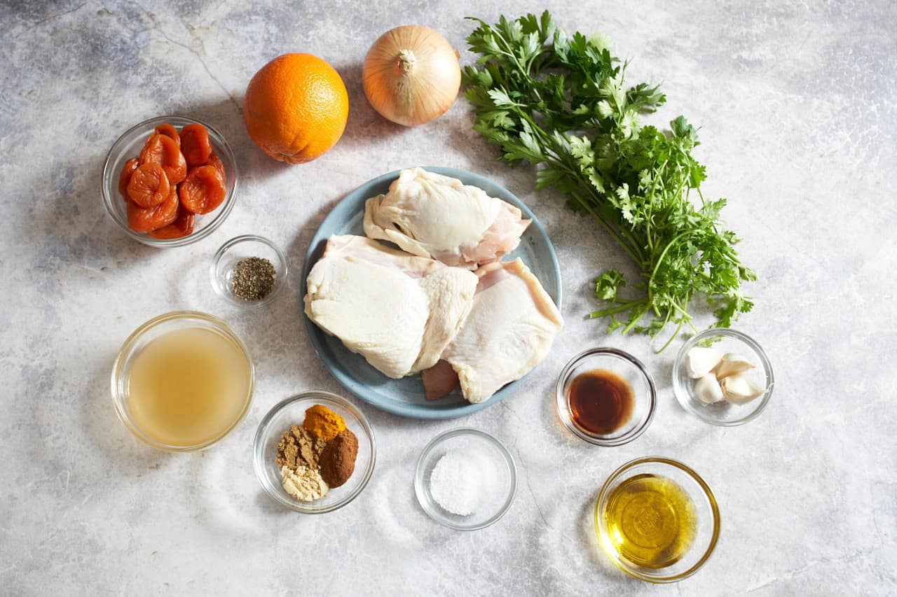 All ingredients for chicken and apricot tagine arranged on a light stone kitchen counter, including chicken thighs, onions, garlic, spices, dried apricots, olives, lemons, and broth