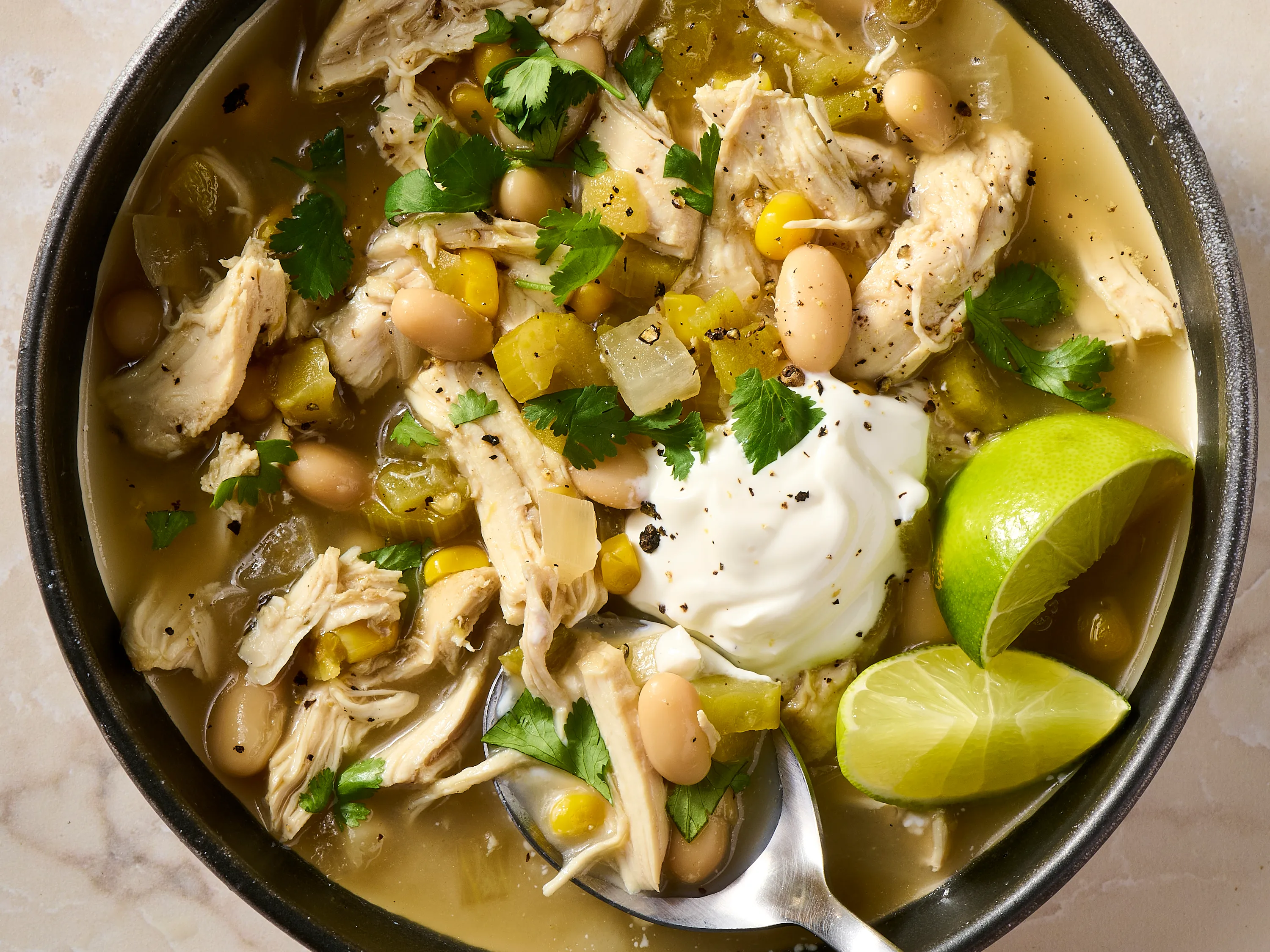 All ingredients for crock pot white chicken chili arranged on a kitchen counter, including chicken breasts, white beans, corn, green chiles, onion, garlic, broth, cream cheese, and spices