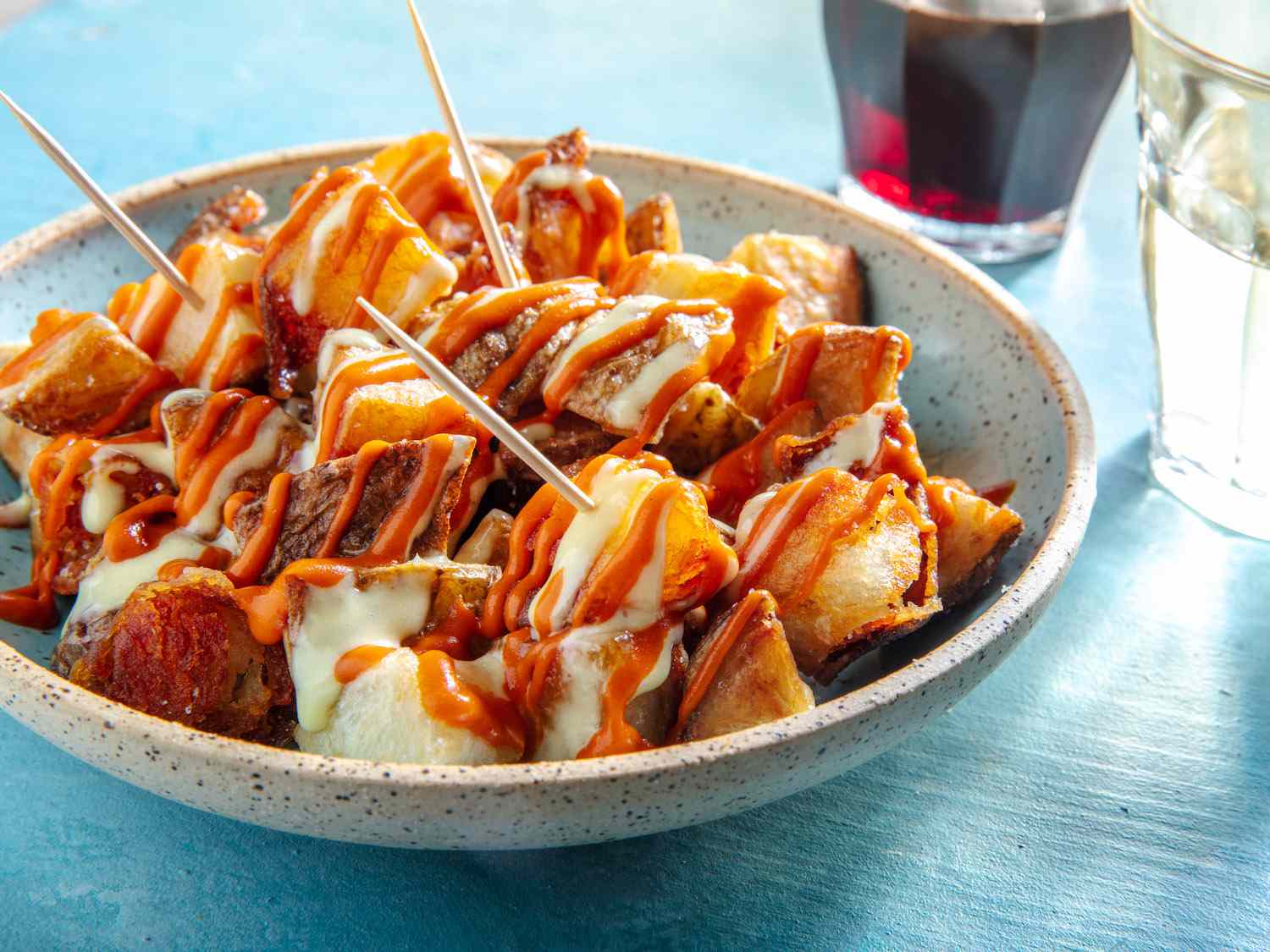 All ingredients for patatas bravas arranged on a bright kitchen counter, including potatoes, olive oil, smoked paprika, garlic, canned tomatoes, mayonnaise, lemon, and spices