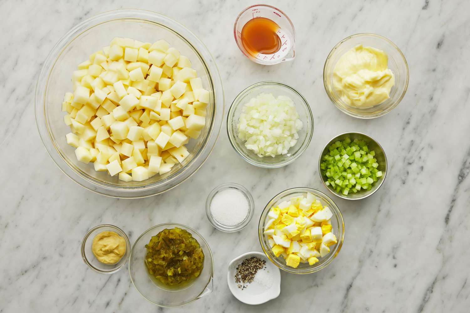 All ingredients for potato salad arranged on a light kitchen counter, including potatoes, mayonnaise, mustard, eggs, celery, red onion, pickles, and herbs