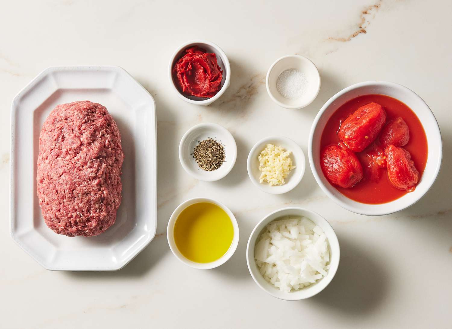 All ingredients for spaghetti meat sauce arranged on a kitchen counter, including ground beef, canned tomatoes, tomato paste, onion, garlic, olive oil, dried Italian seasoning, salt, pepper, and a small bowl of grated Parmesan