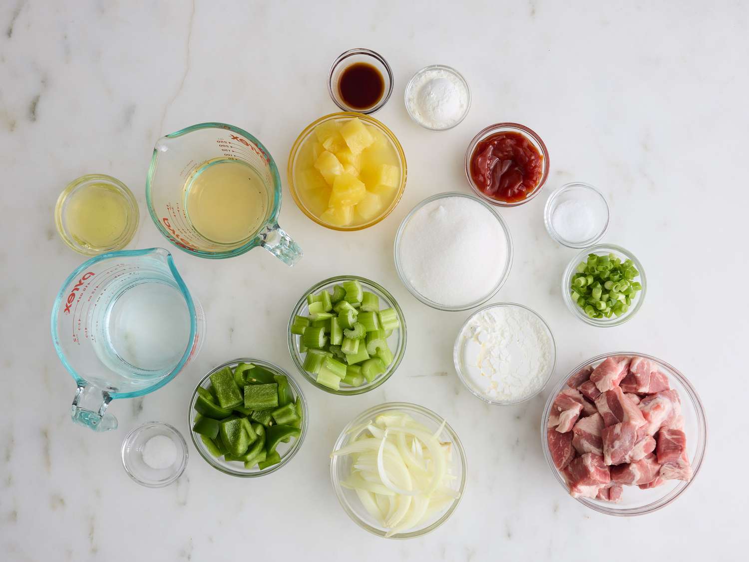 All ingredients for sweet and sour pork arranged on a kitchen counter: cubed pork, pineapple chunks, bell peppers, onion, garlic, ginger, ketchup, rice vinegar, soy sauce, brown sugar, cornstarch, eggs, and neutral oil