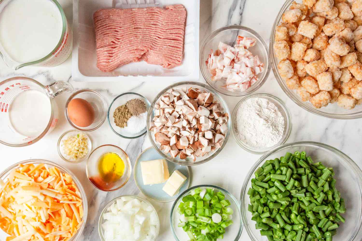 All ingredients for tater tot hotdish arranged on a kitchen counter including frozen tater tots, ground beef, onion, garlic, cream of mushroom soup, sour cream, cheddar cheese, and frozen mixed vegetables