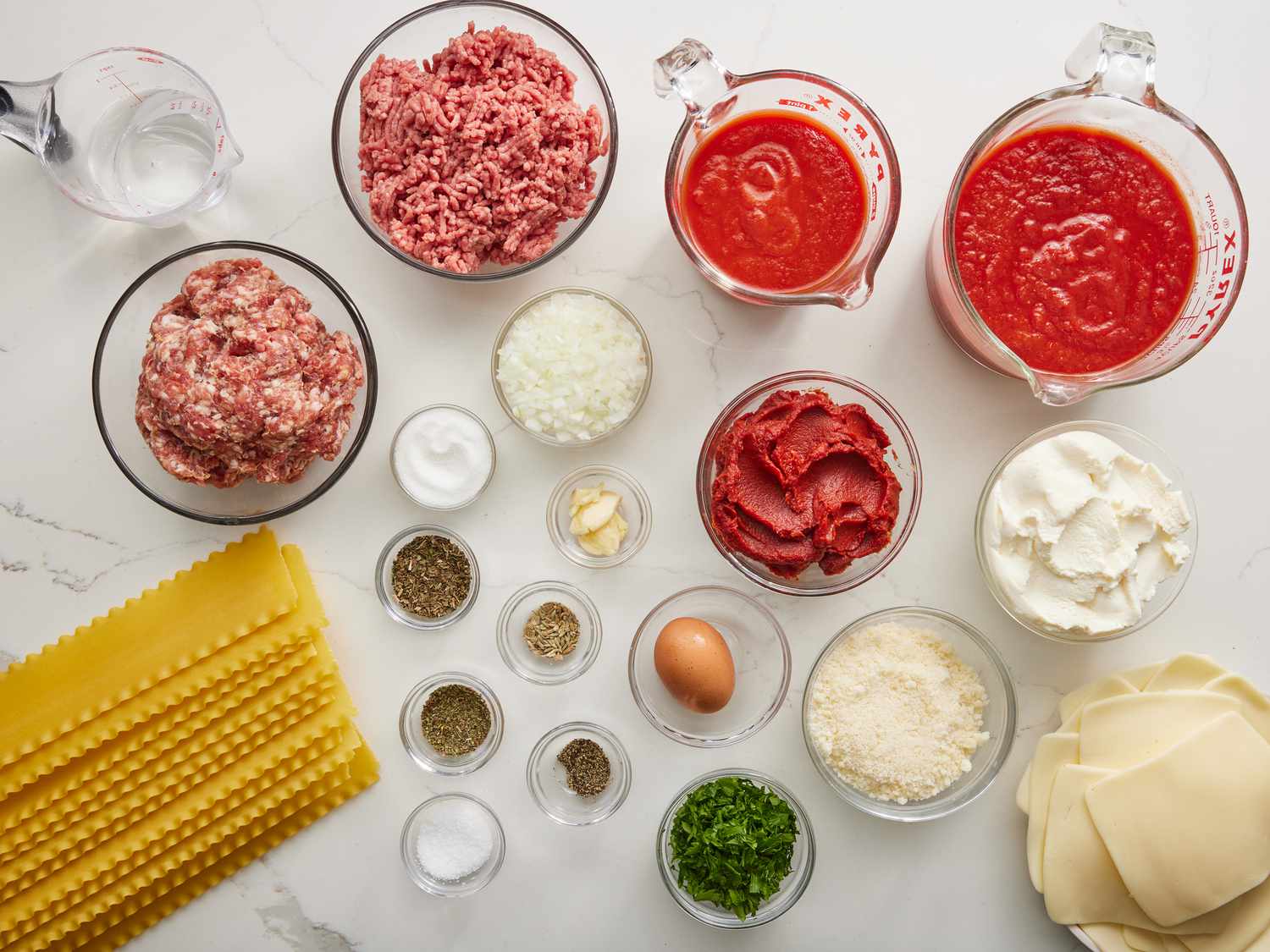All lasagna ingredients arranged on a clean kitchen counter, including ground beef, Italian sausage, canned tomatoes, ricotta, mozzarella, Parmesan, lasagna noodles, onions, garlic, and herbs in small bowls