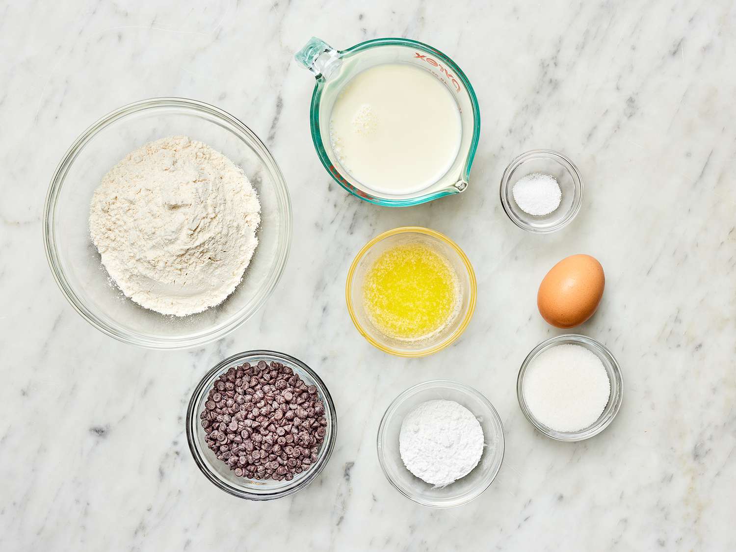 All-purpose flour, sugar, baking powder, baking soda, salt, eggs, milk, melted butter, vanilla, and chocolate chips arranged on a bright kitchen counter