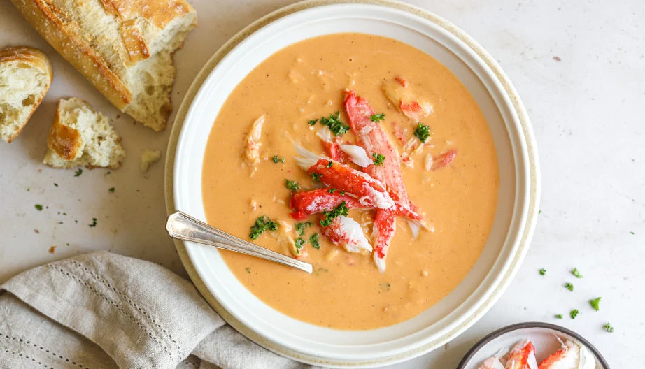 All the crab bisque ingredients arranged on a clean kitchen counter including crab meat, onion, celery, garlic, tomato paste, cream, broth, and herbs in small bowls