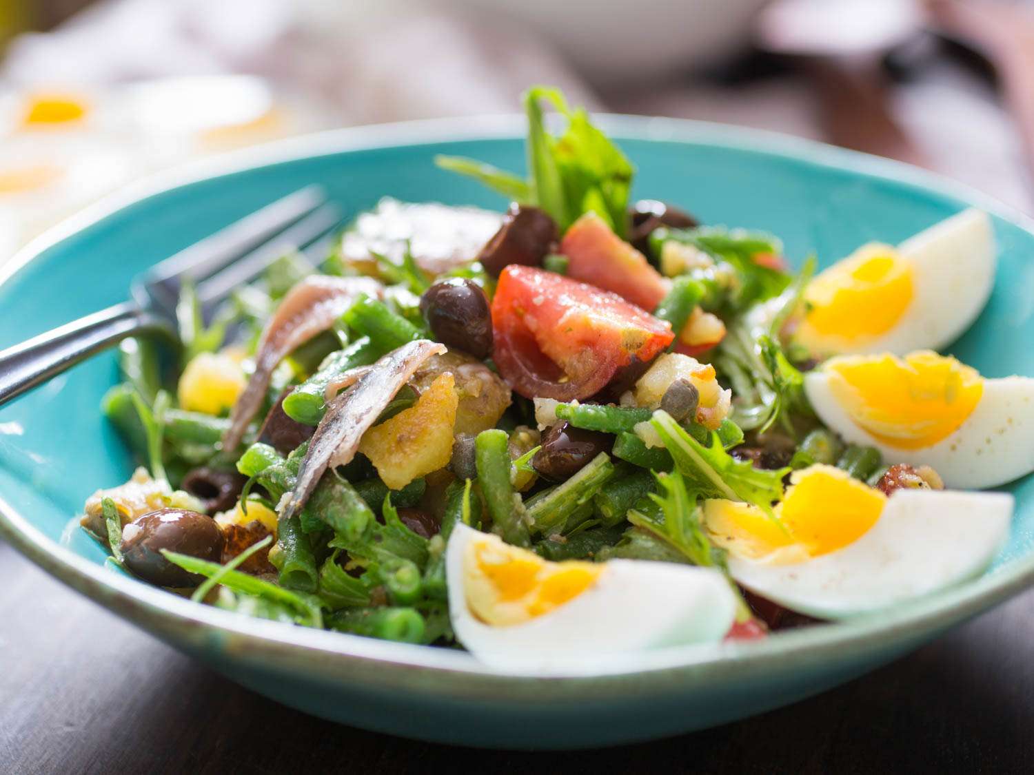 All the ingredients for Salad Niçoise laid out on a clean kitchen counter including eggs, baby potatoes, green beans, tomatoes, olives, tuna, Dijon mustard, lemon, and herbs in small bowls