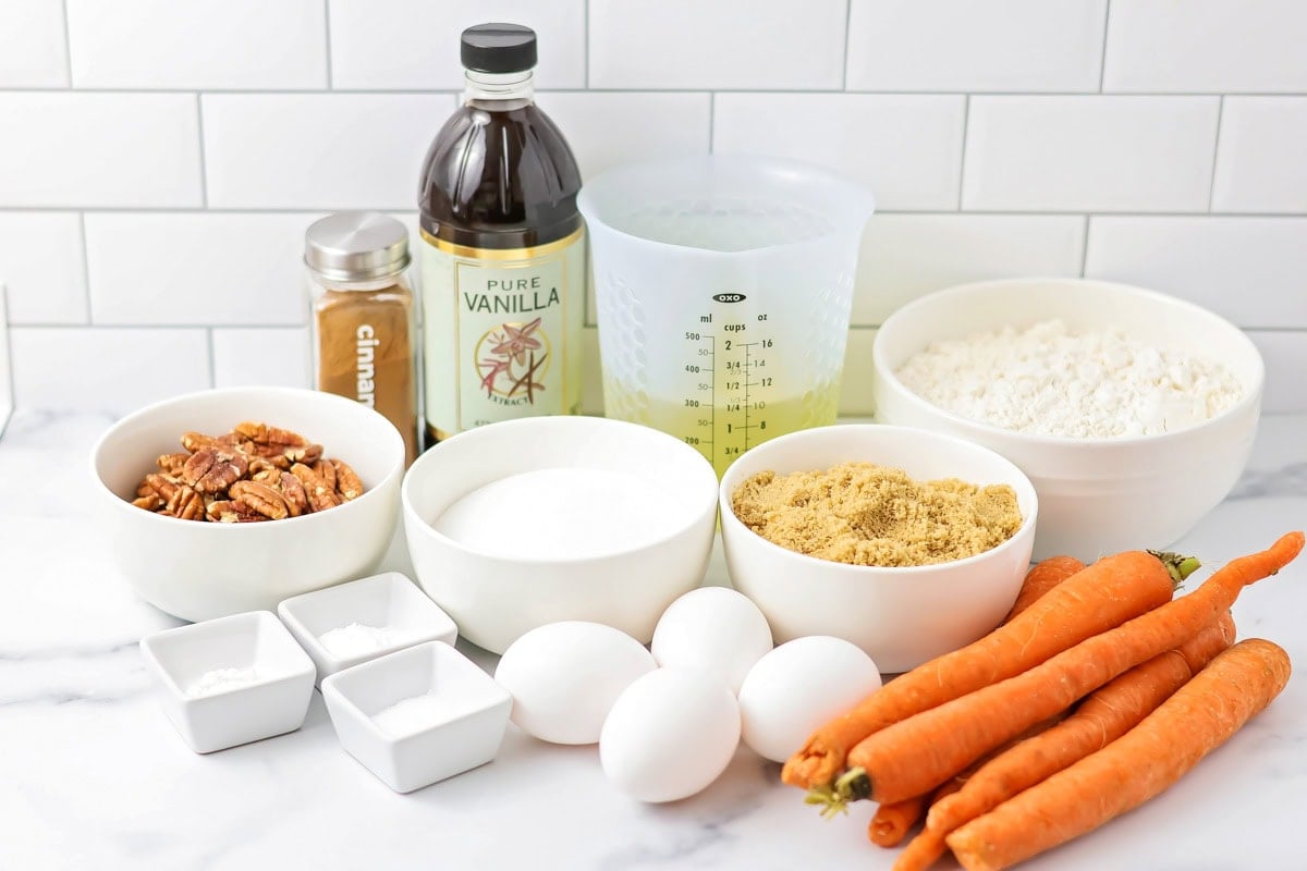 All the ingredients for carrot cake arranged on a tidy kitchen counter, including grated carrots, flour, sugar, eggs, oil, spices, and cream cheese