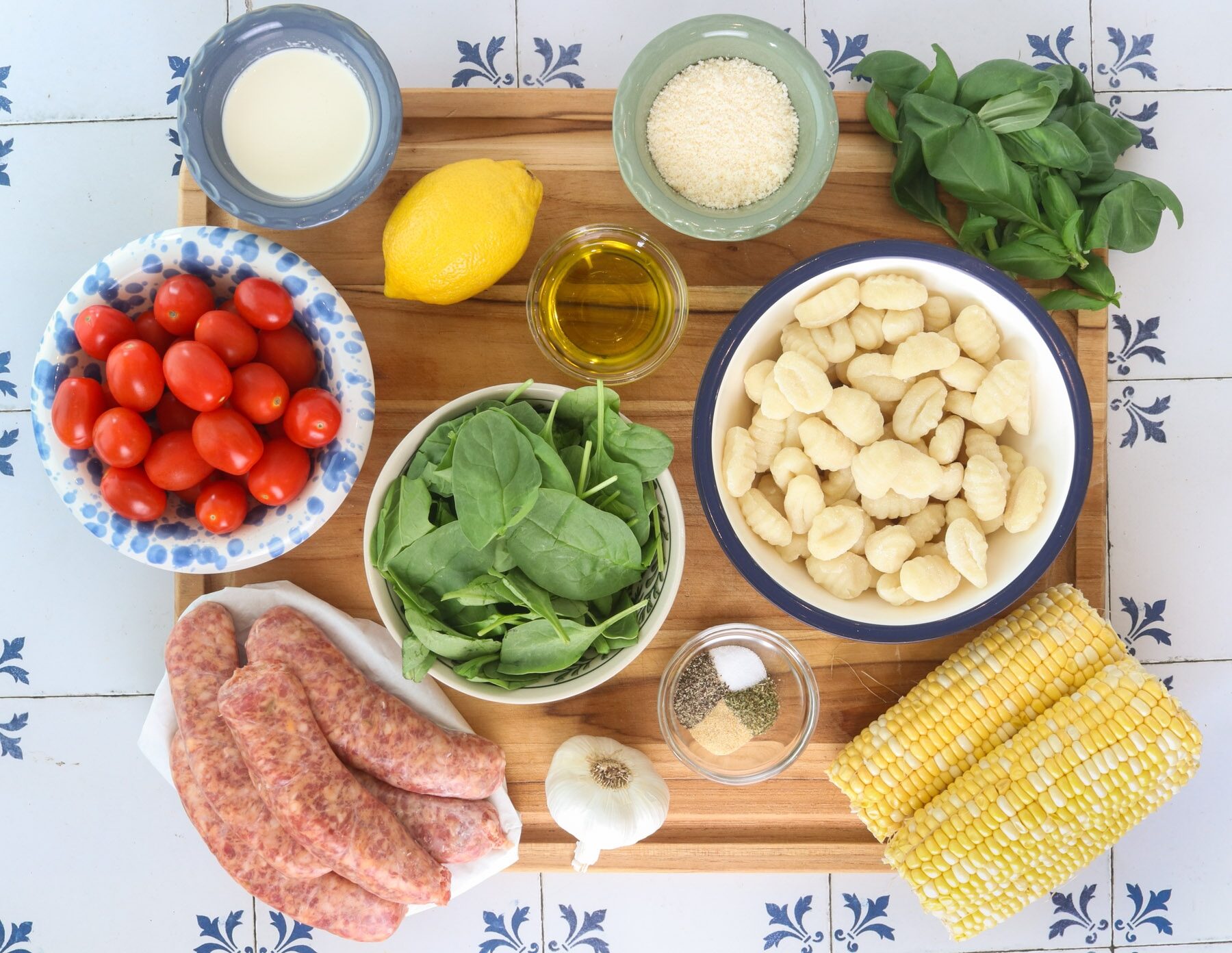 All the ingredients for sheet pan gnocchi laid out on a light kitchen counter, including packaged potato gnocchi, cherry tomatoes, zucchini, red onion, garlic, lemons, basil, Parmesan, olive oil, and spices