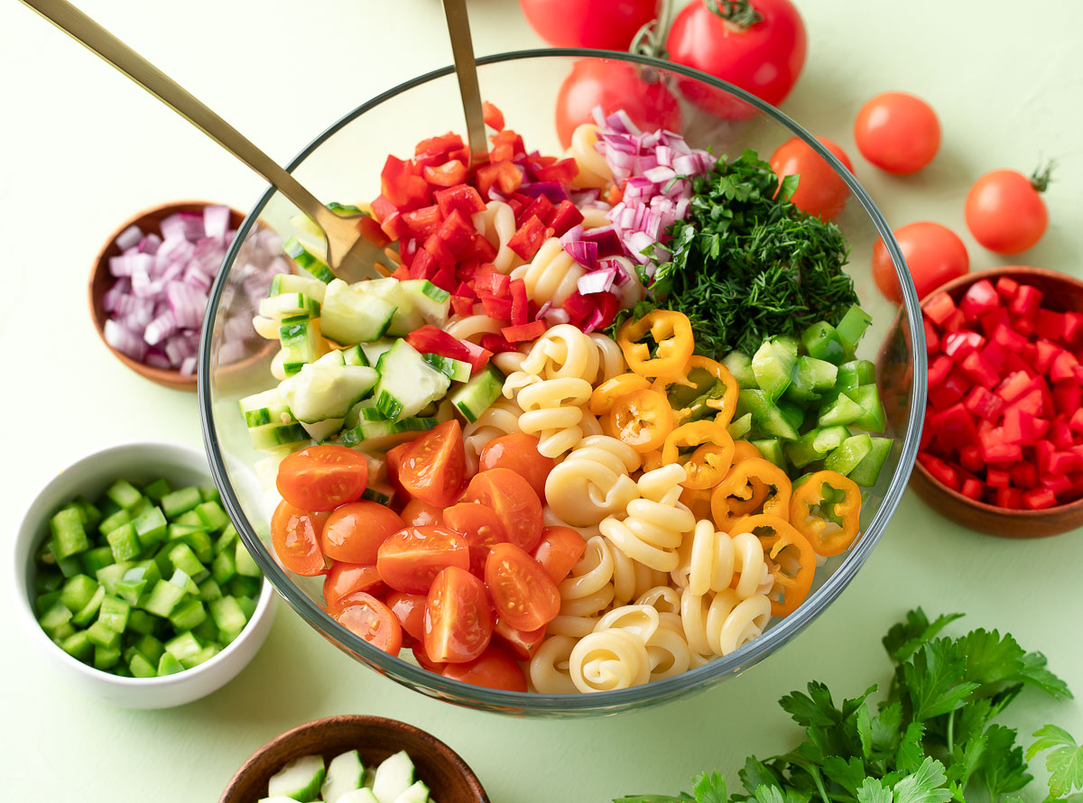 All the pasta salad ingredients arranged on a kitchen counter including rotini, cherry tomatoes, cucumber, red onion, feta, pepperoncini, olive oil, vinegar, and dried herbs